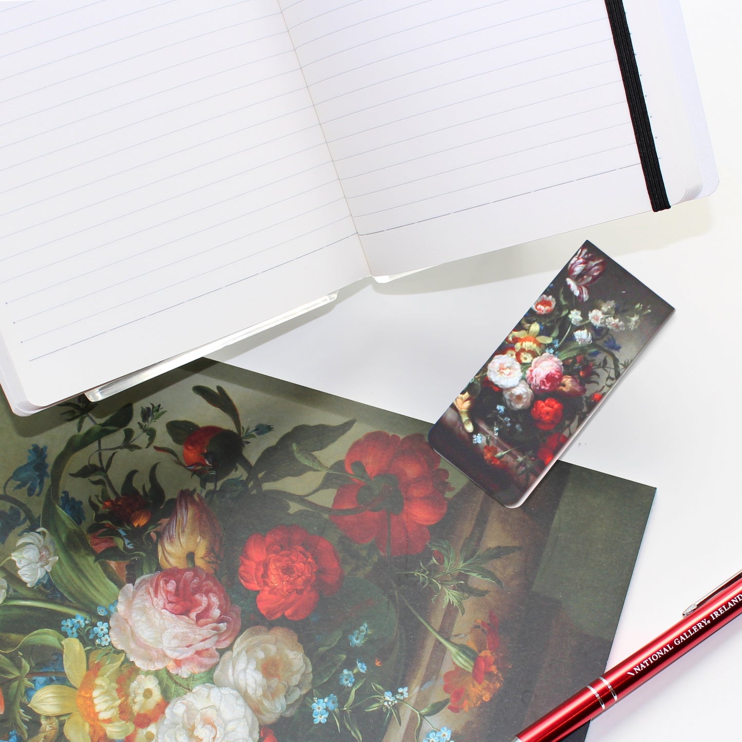 A sketchbook, magnetic bookmark and open notebook featuring Rachel Ruysch's Vase of Flowers with and Ear of Corn still-life painting on a white background, accompanied by a red National Gallery pen.