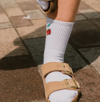 A close-up of a person's foot wearing a white ribbed crew sock with a small red cherry graphic, paired with tan double-strap sandals on a paved outdoor walkway.
