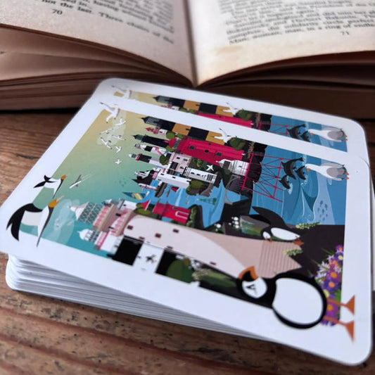 A deck of cards lies face down on a wooden table. Their backs show an illustrated scene of Irish Lighthouses. 
