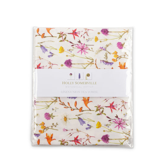 A clear pack with a belly band of the designer’s name and logo around the centre. A folded towel is visible inside, covered in a pattern of wildflowers in orange, yellow, red, blue, pink, and purple.