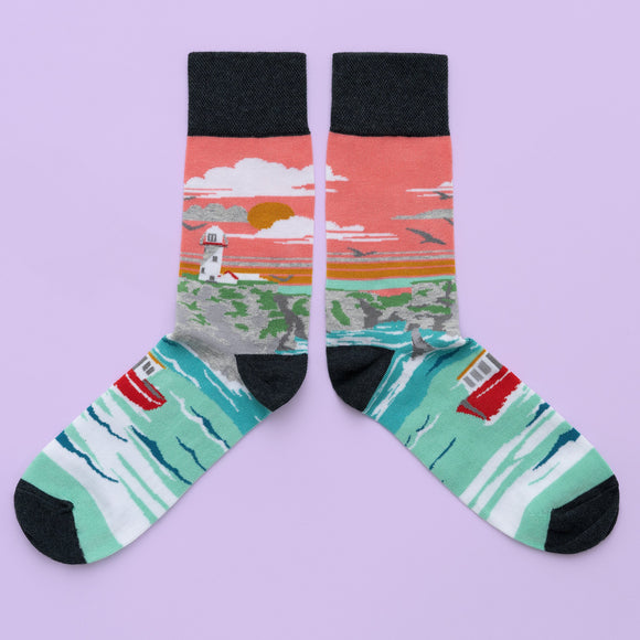 A pair of socks with a white lighthouse on a cliff around the leg. A peach sky with grey birds is in the background. The foot is a red boat on a light blue sea, with a black toe, heel and cuff.