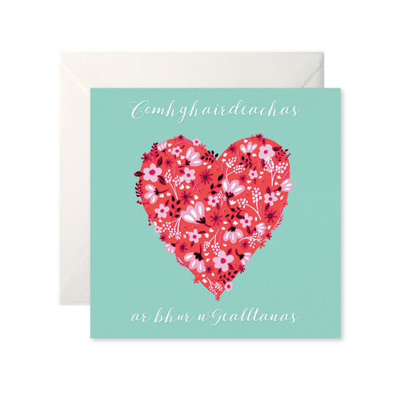 A mint card with a large red heart made up of flowers in the centre. Above is written ‘Comhghairdeachas’ and below is ‘ar bhur nGealltanas’ both in white cursive.