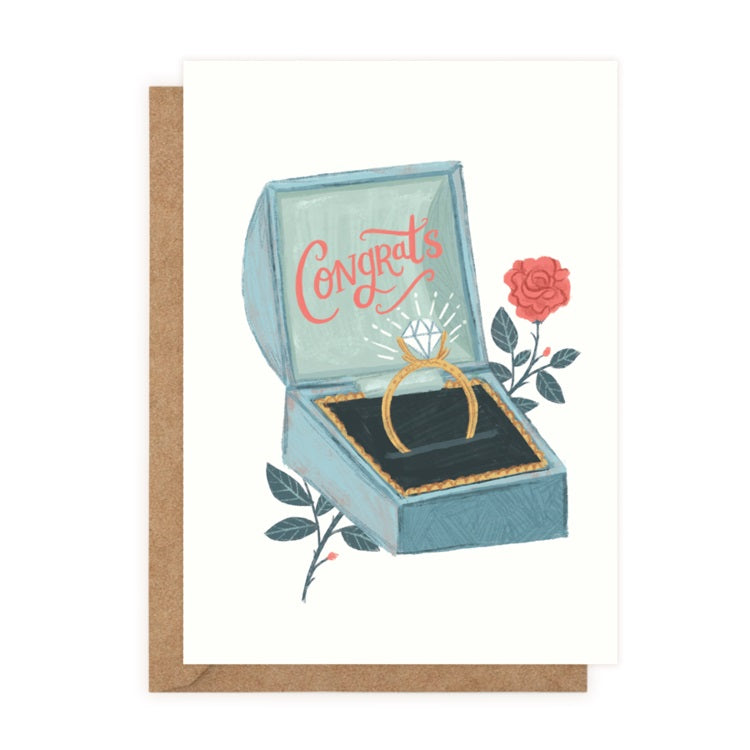 A white card with a drawing of an open blue ring box. Inside the lid ‘Congrats’ is written in red cursive. In the base is a gold diamond ring. A red rose is behind the box.