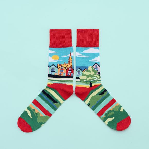 A pair of socks with a landscape of colourful houses around the leg. A cathedral, blue sky and the sea are in the background. There are matching coloured stripes along the foot, with a red toe, heel and cuff.
