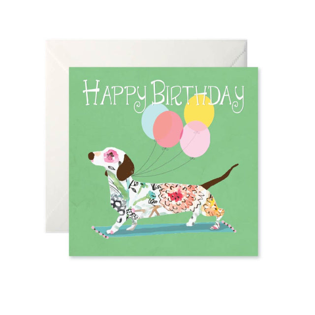 A green card with ‘Happy Birthday’ written in white across the top. Below is a white dog covered in a floral pattern with four balloons attached to it.