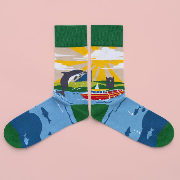 A pair of socks with a seascape of a dolphin jumping out of the water. A boat, landscape and yellow sky are in the background. The foot is blue water with the outline of fish, with a dark green toe, heel and cuff.