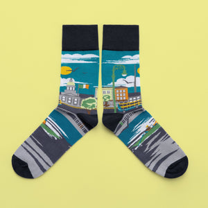 A pair of socks with a cityscape around the leg. A buildings, street lamps, a bus and blue sky are in the background. The foot is green canoe on the river, with a black toe, heel and cuff.
