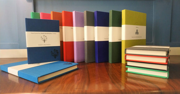 A selection of clothbound notebooks, standing and stacked, each a different colour with a cream belly band around the centre. There is a simple line illustration of a street lamp with five lanterns on the band and in the bottom right corner of the notebooks.