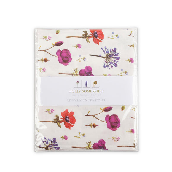 A clear pack with a belly band of the designer’s name and logo around the centre. A folded towel is visible inside, covered in a pattern of wildflowers in red, blue, pinks, and purples.
