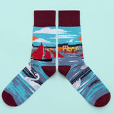 A pair of socks with a red sailed boat on the sea around the leg. Colourful buildings and a sunny sky are in the background. The foot is swans on the water, with a dark wine toe, heel and cuff.