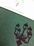 A close up of a lamp illustration on a dark green cloth notebook cover. A matching cream belly band is visible.