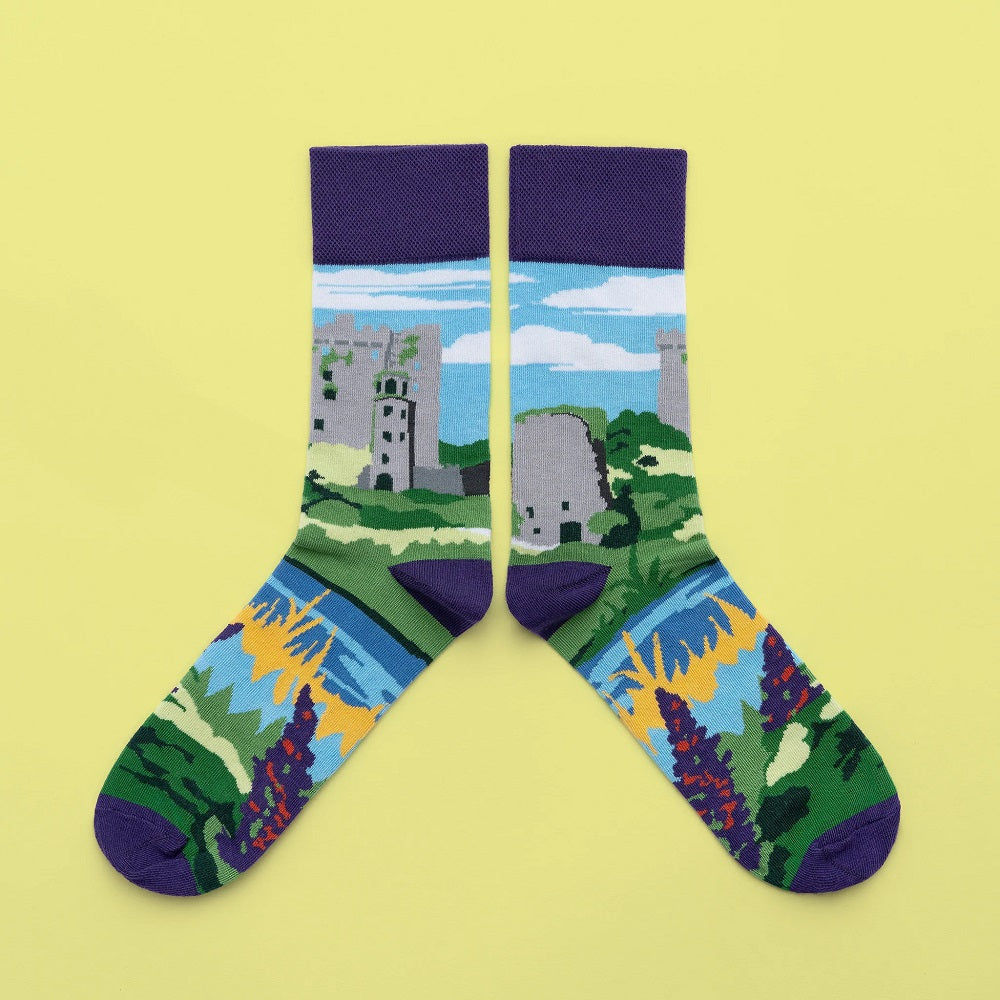 Socks with a castle in a rural landscape with blue skies around the leg, a river and purple flowers around the foot. 