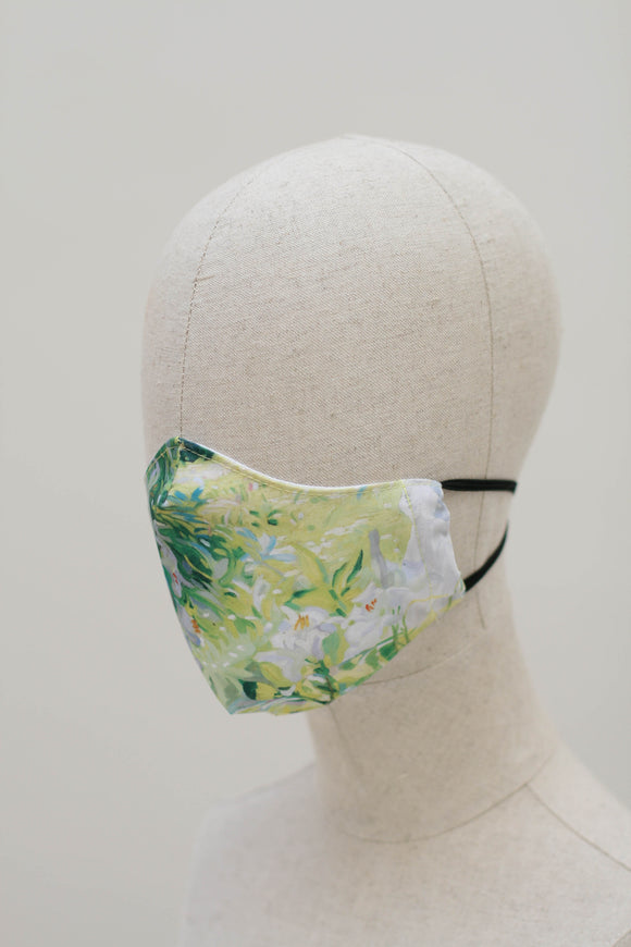 A shaped face mask on a mannequin head. The design is a painting of white flowers on a background of green made up of grass and leaves. It has black ear straps.
