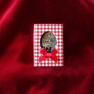 A gold coloured pin of a flower bouquet inside a red and white diamond pattern box with a small ribbon bow.