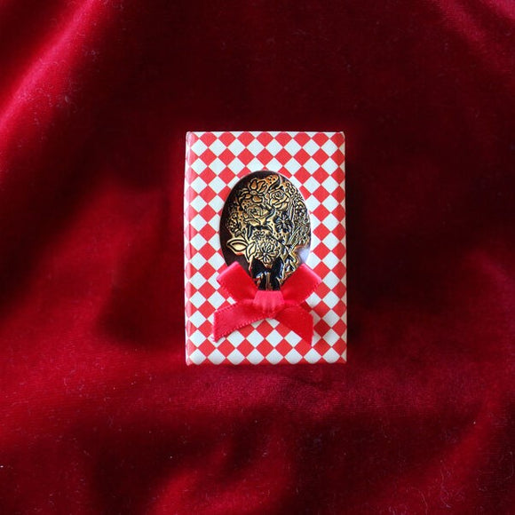 A gold coloured pin of a flower bouquet inside a red and white diamond pattern box with a small ribbon bow.