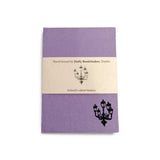 A light purple clothbound notebook with a cream belly band around the centre. There is a simple line illustration of a street lamp with five lanterns in the bottom right corner of the notebook and on the band.