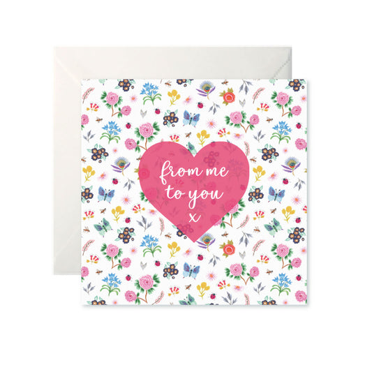 A white card with a pattern of colourful, small, simple flowers and butterflies. A pink heart is in the centre with ‘from me to you x’ in white cursive inside.