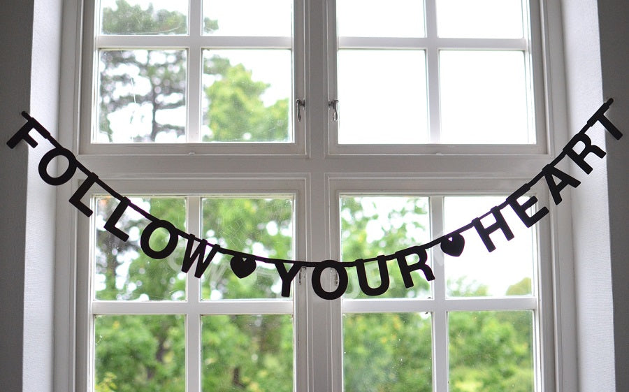 A banner of black letters in a window spelling Follow Your Heart with little cut out hearts in between the words.