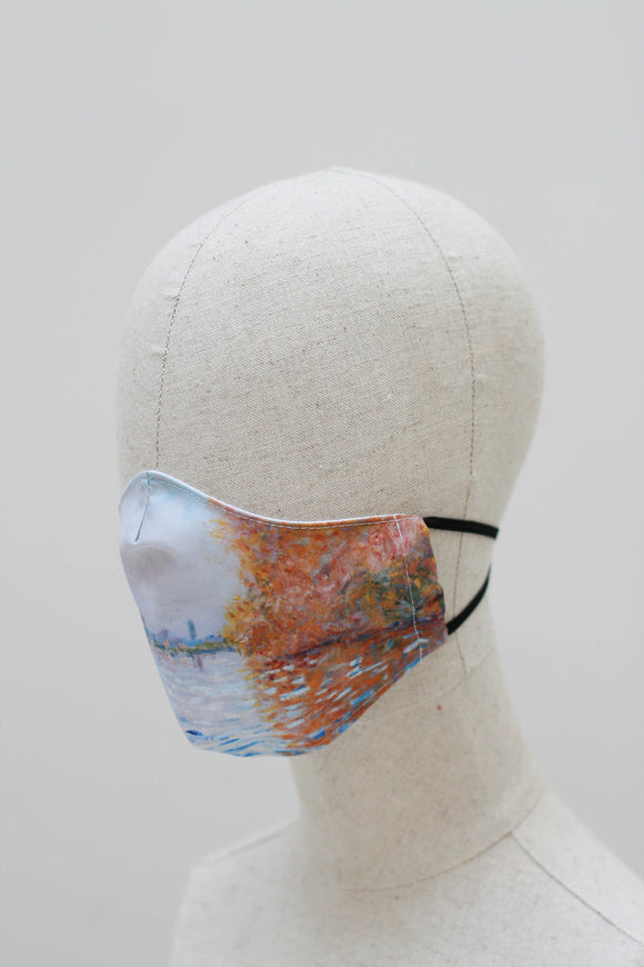 A shaped face mask on a mannequin head from a side angle. The design is an impressionist style painting of an orange autumn tree reflected into a light blue lake, with a matching light blue sky. The lake and sky continue around the mask. It has black ear straps.