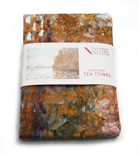 A folded tea towel with a grey label around the centre. The label shows aan impressionist painting of a light blue sky and matching coloured lake. The right of the image is dominated by a large tree in autumn oranges which reflects into the water. On the left is a small boat with its white sail open.