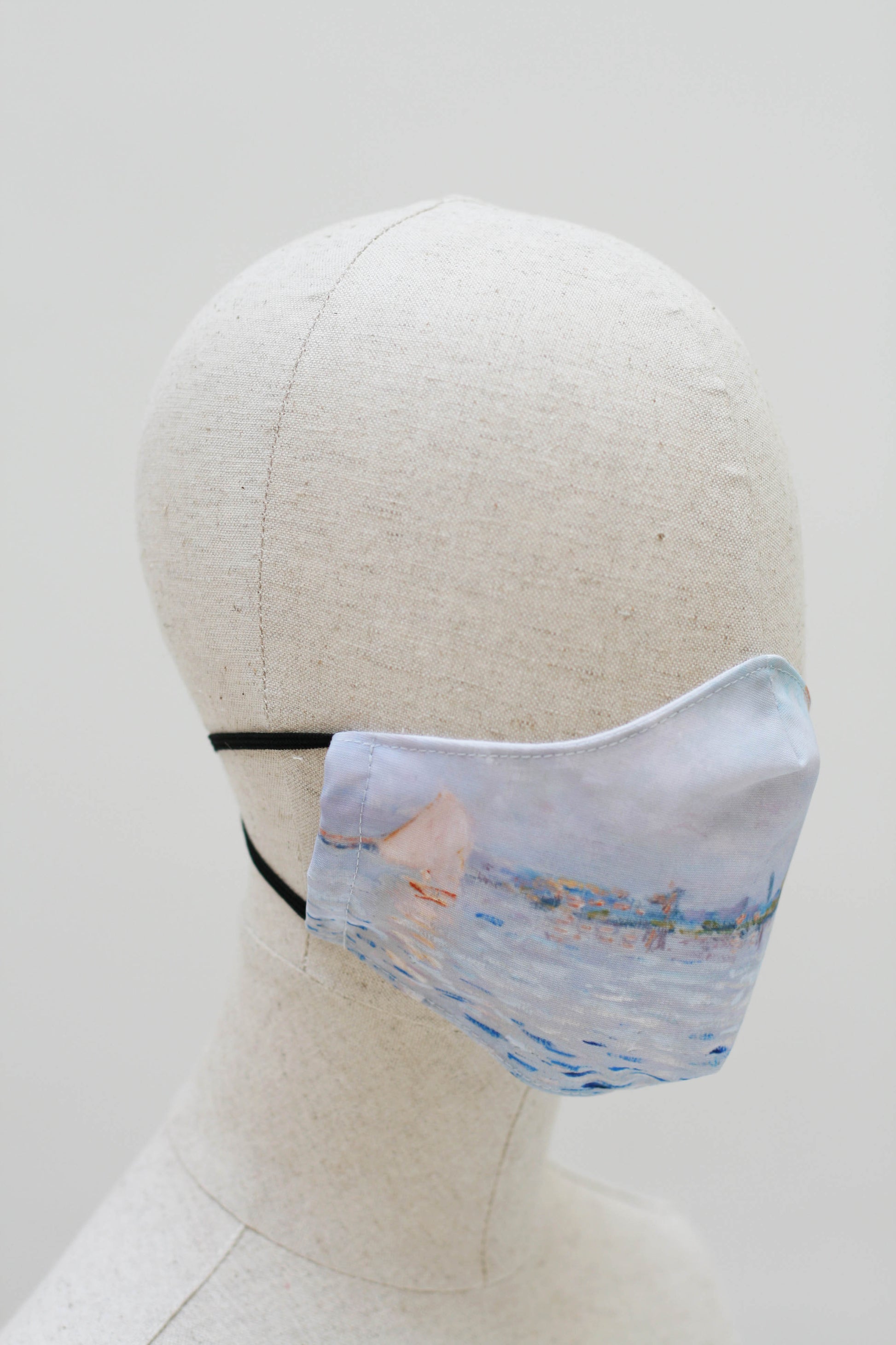 The mask on a mannequin head from the other side. The lake and sky continue with a single sailboat, its white sails open, towards the edge of the mask.