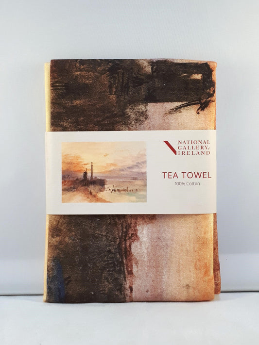 A folded tea towel with a grey label around the centre. The label shows a vibrant watercolour of a beachside scene at sunset. A lighthouse and windmill are silhouetted in black against an orange and purple sky.