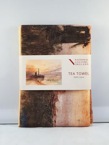 A folded tea towel with a grey label around the centre. The label shows a vibrant watercolour of a beachside scene at sunset. A lighthouse and windmill are silhouetted in black against an orange and purple sky.