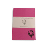 A pink clothbound notebook with a cream belly band around the centre. There is a simple line illustration of a street lamp with five lanterns in the bottom right corner of the notebook and on the band.
