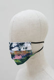 A pleated face mask on a mannequin head. The design is of an impressionist style landscape at the edge of a forest in shades of green, pink, brown and an almost white sky. It has black ear straos.