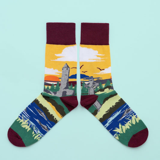 A pair of socks with a landscape with a round tower and a large stone Celtic cross around the leg. A forest. setting sun and yellow sky is in the background. The foot is a river with grassy banks, with a wine toe, heel and cuff.