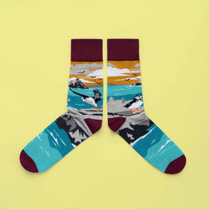 A pair of socks with puffins on rocks in a seascape. A mountain island (Skellig), flying birds and a mustard sky are in the background. The foot is more of the sea, with a wine toe, heel and cuff.