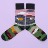 A pair of socks of a rural landscape with thatched cottages and a sheep. Mountains, water and a pink sky are in the background. The foot is more fields and a sheep, with a wine toe, heel and cuff.
