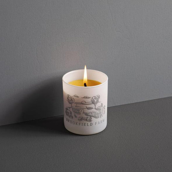 A lit candle in a semi-opaque white jar with a grey illustration of an idealised farm with trees, sheep, a tractor and a setting sun in the background. The angle shows the yellow wax inside.