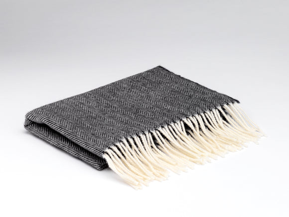 A folded dark grey wide scarf with white fringe on the ends. There is a herringbone pattern across the wrap.
